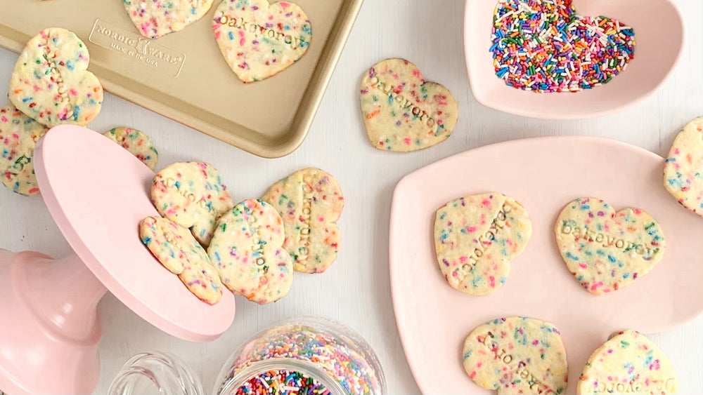 Bakeyorey Funfetti heart shape cookies on a gold baking sheet, pink cookie plate and rainbow sprinkles in a pink heart shaped bowl