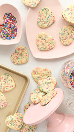 Bakeyorey Funfetti heart shape cookies on a gold baking sheet, pink cookie plate and rainbow sprinkles in a pink heart shaped bowl
